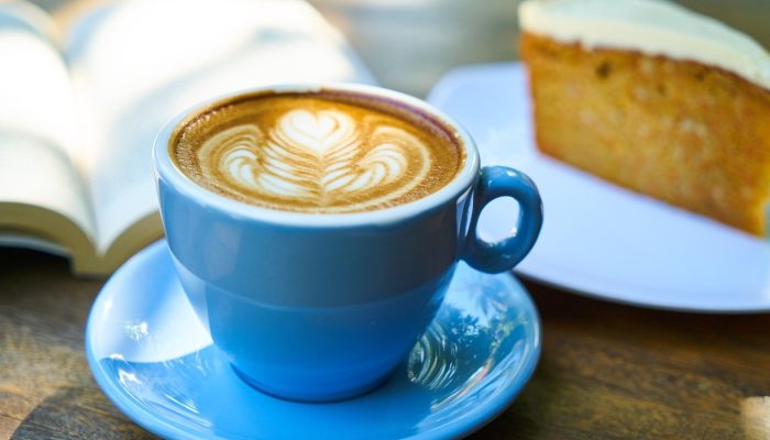 Cappuccino Latte Art blauen Tasse mit Untertasse, ein Stück Karottenkuchen mit einer Frischkäse Glasur und ein aufgeschlagenes Buch auf einem Holztisch, Sonnenlicht wirft Schatten auf die Nahaufnahme
